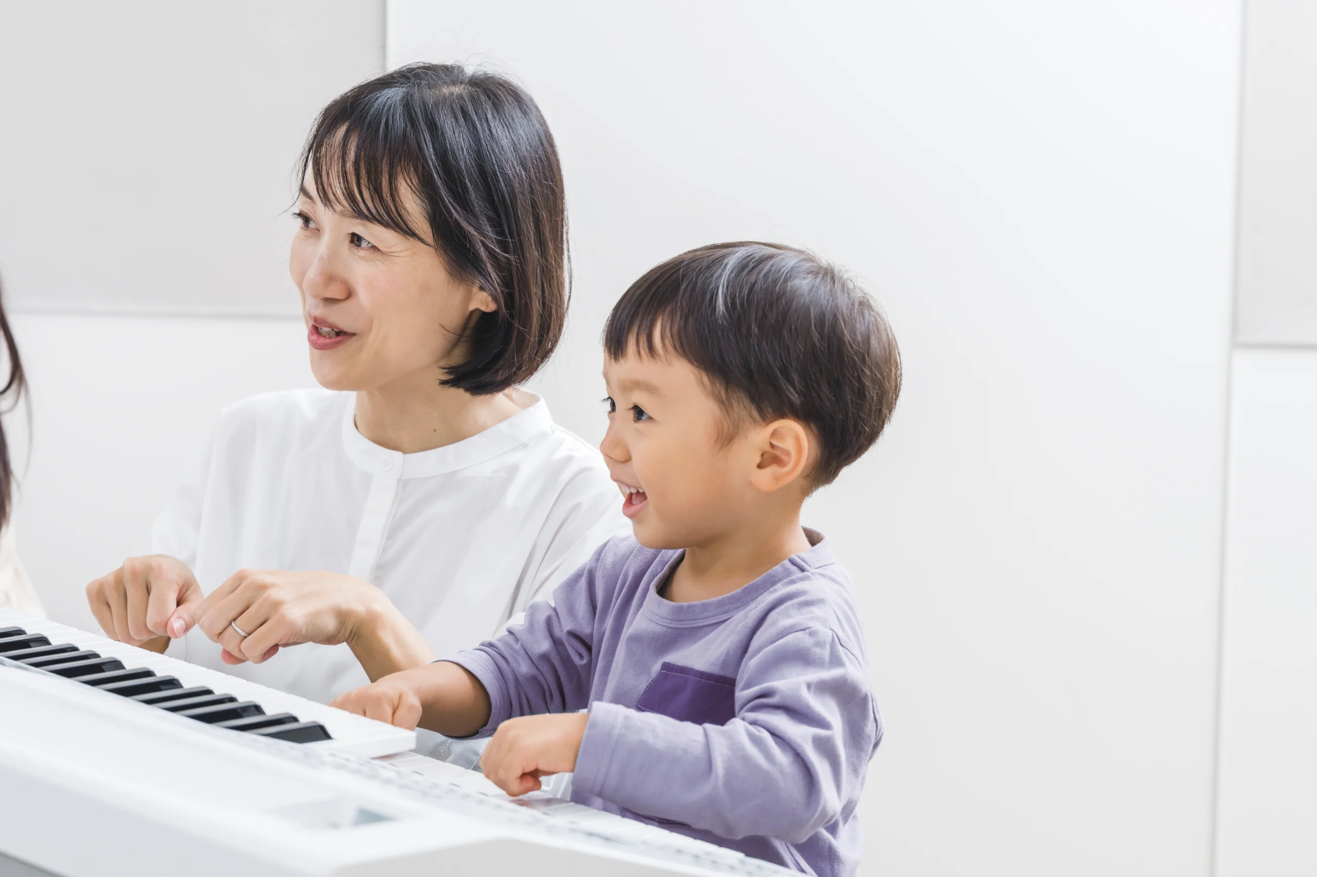 A smiling young child learning to play keyboard with a supportive female teacher at Yamaha Music School – joyful early music education for children.