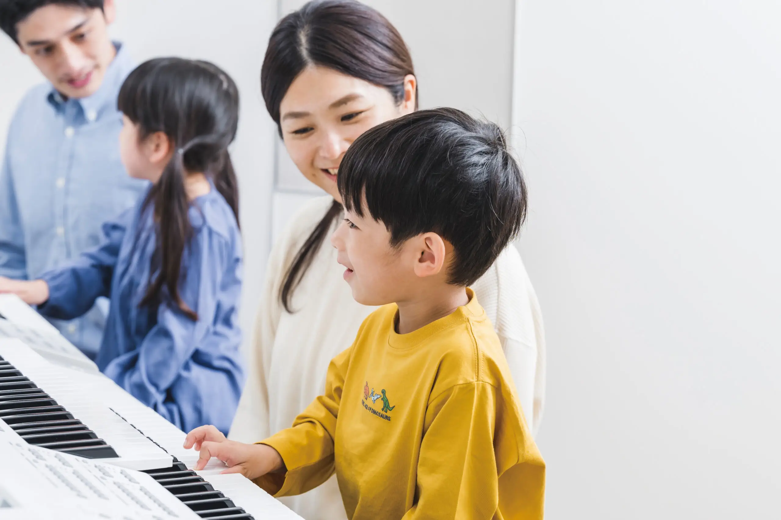 Young child in yellow jumper playing piano with mother sitting beside him, while another child plays electone with father in the background at Yamaha music school